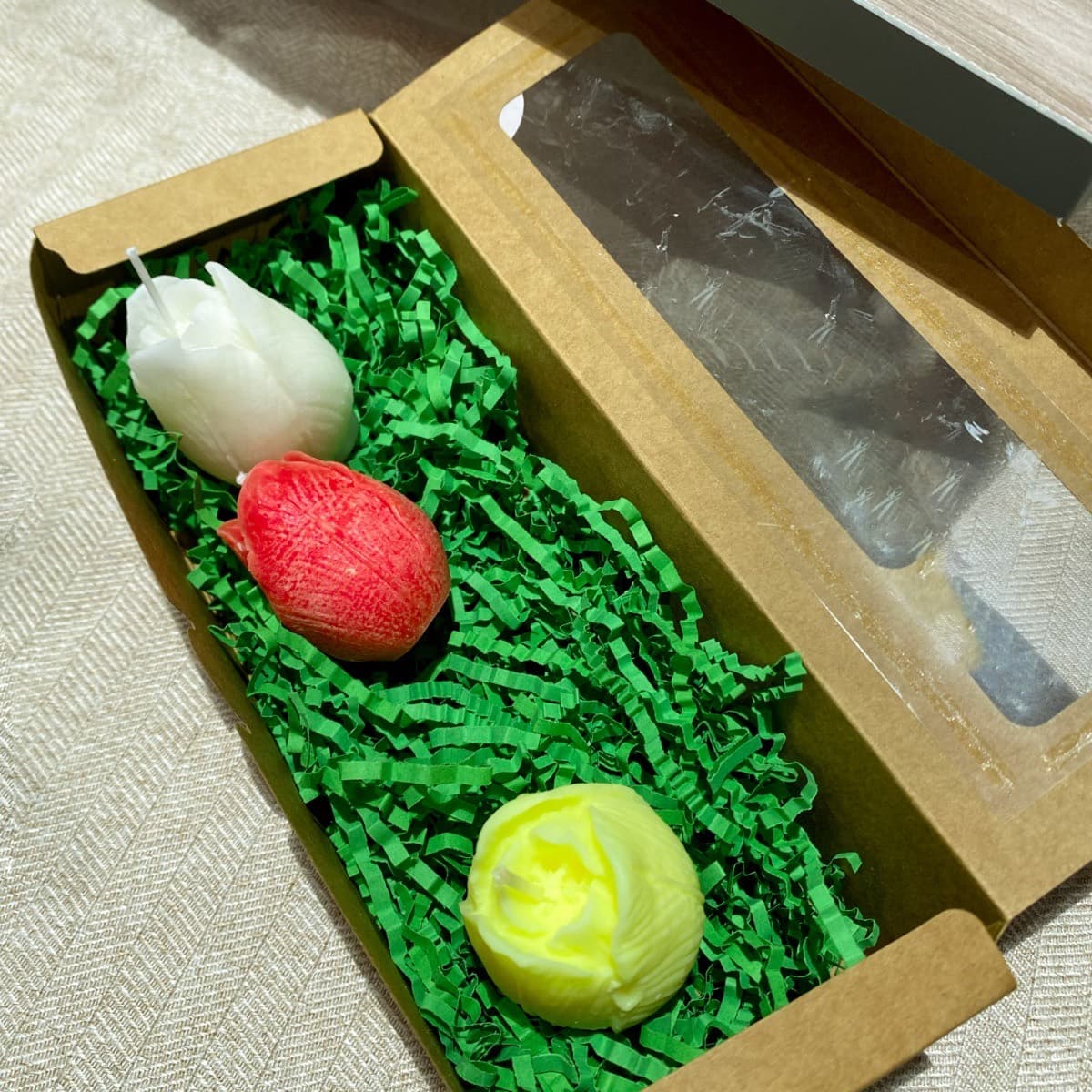 Three tulip candles — white, red and yellow — in a kraft gift box on green paper grass